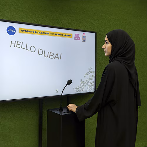 Arab woman using Sayit AI at a Dubai corporate event, speaking into a mic as 3D branded text appears on a large screen, showcasing interactive event technology rental