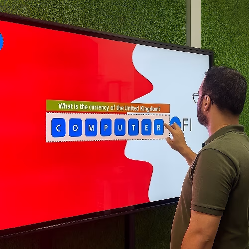 Guest playing word scramble game on large touchscreen at Dubai event with branded graphics and green artificial grass wall background.