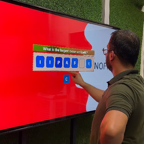 Guest playing word scramble game on large touchscreen at Dubai event with branded graphics and green artificial grass wall background.