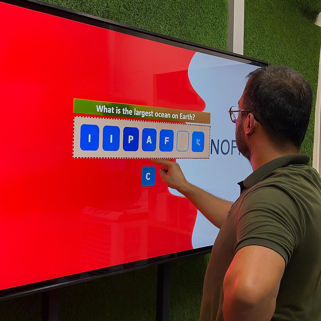 Guest playing word scramble game on large touchscreen at Dubai event with branded graphics and green artificial grass wall background.